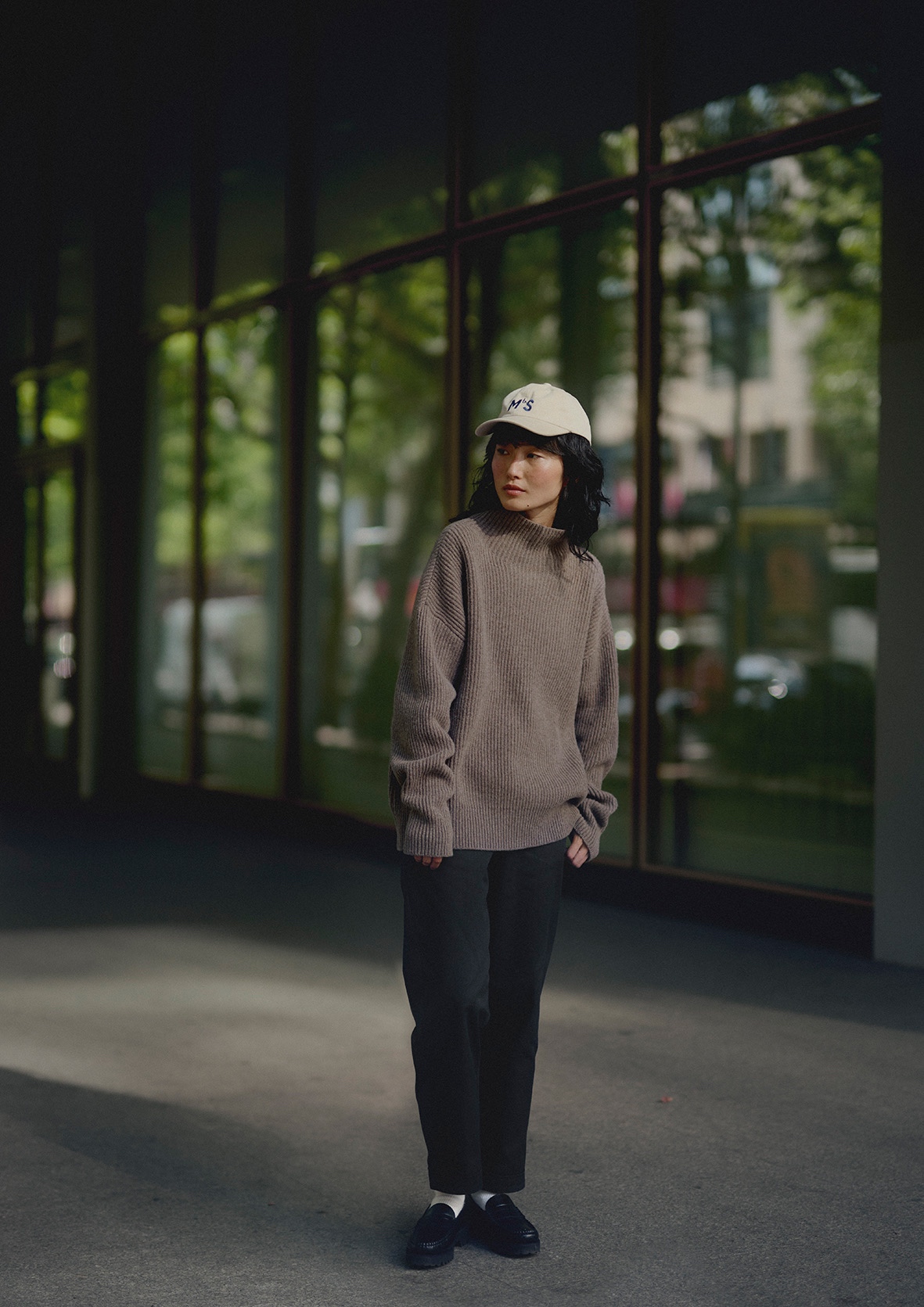 Woman wearing grey turtleneck pullover, black pants and white cap in front of a window
